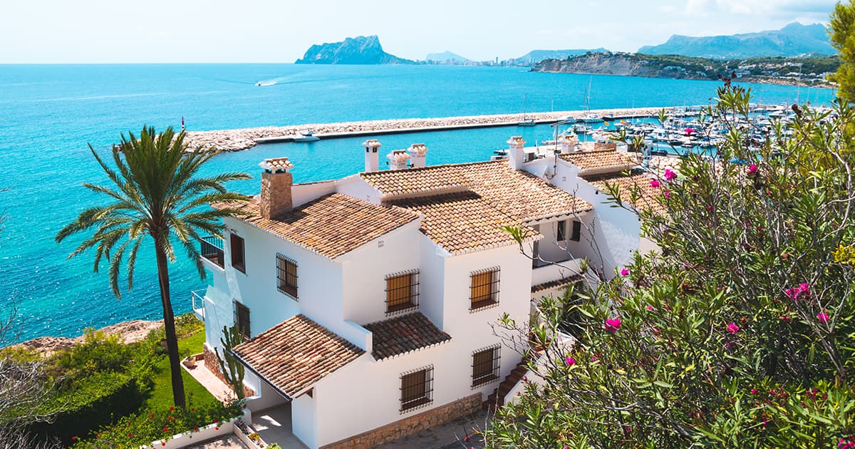 White seaside house with terracotta roof, palm tree, overlooking a marina and blue sea.